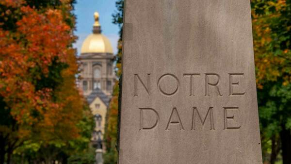 The main entrance to Notre Dame campus is pictured with the Golden Dome / Main Building in the background and the words Notre Dame carved into stone in the foreground.