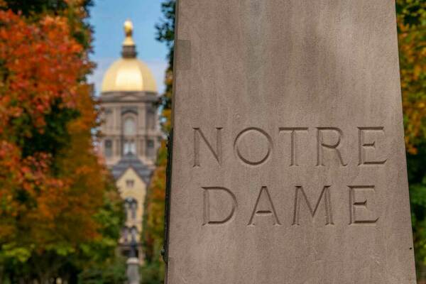 The main entrance to Notre Dame campus is pictured with the Golden Dome / Main Building in the background and the words Notre Dame carved into stone in the foreground.