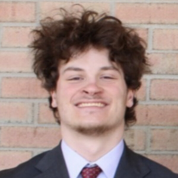 Nathan Barr. A young man with curly brown hair, wearing a dark suit, light purple shirt, and red tie.